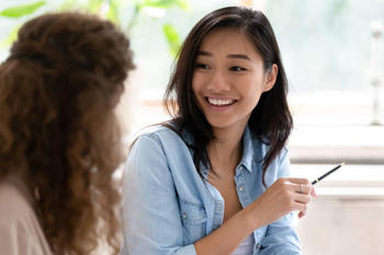 Woman in button down smiling and pointing at something with a pen to another woman, whose face is turned away from the camera.