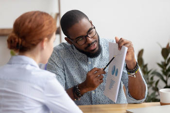 Man pointing to something on a document with graphs and text on it, to a woman sitting beside him.